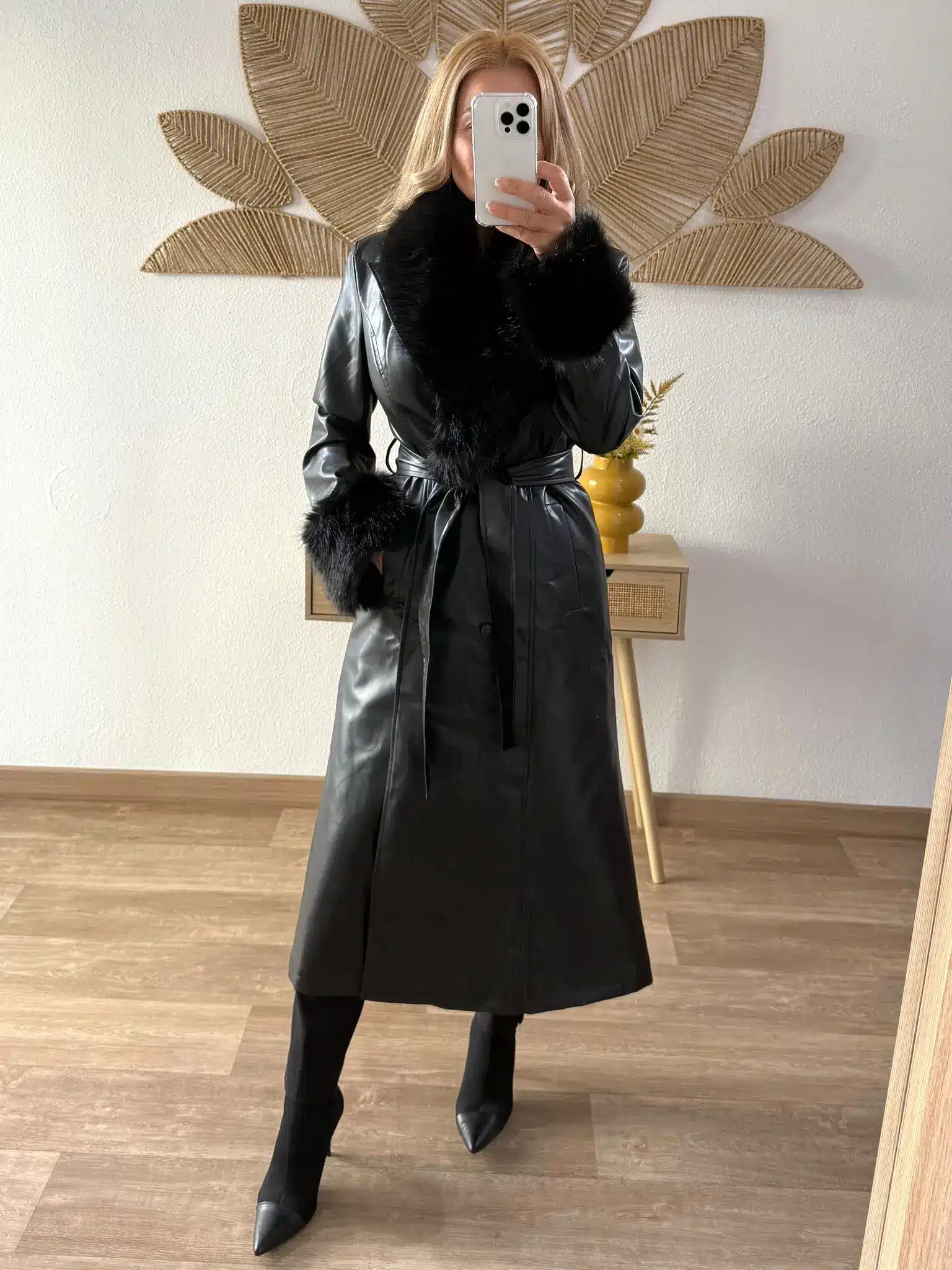 Abrigo-polipiel-cuello-y-puños-pelo-1 Mujer usando un abrigo largo de polipiel negro con cuello y puños de pelo sintético, combinado con botas negras de tacón.