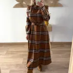 Mujer con vestido largo de cuadros en tonos marrones y beige, de corte recto y cinturón a juego. Lleva botas de ante marrón claro con punta en forma de pico.