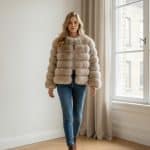 Mujer caminando en interior luminoso con abrigo corto de pelo voluminoso color beige, jeans ajustados azules y botines marrones.