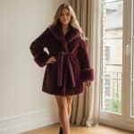 Abrigo de pelo sintético color burdeos, de largo medio muslo (por debajo de los glúteos), con cinturón a la cintura, cuello amplio y puños voluminosos.