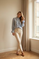 Mujer con camisa azul claro de manga larga con volantes frontales y cuello alto, combinada con pantalón beige y zapatillas blancas.