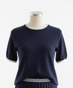 Mujer caminando en interior luminoso con conjunto beige: camiseta de punto con ribete marrón, falda plisada corta y zapatillas blancas.