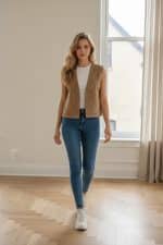 Mujer con camiseta blanca sin mangas y jeans azules usando un chaleco abierto color camel con detalles perforados decorativos, en interior con luz natural.