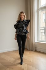 Chaleco negro de estilo vanguardista en material polipiel, con volantes estructurados, cremallera asimétrica y cinturón con hebilla, presentado sobre fondo neutro.