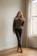 Chaqueta corta de polipiel marrón con cuello de pelo sintético, cierre frontal con botones metálicos y bolsillos con solapa.