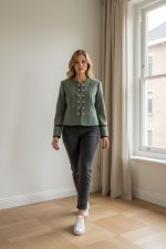 Mujer caminando con chaqueta entallada verde oliva de botones frontales, sin solapas, combinada con pantalón oscuro y zapatillas blancas.