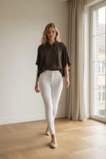 Mujer joven caminando en un interior luminoso junto a una ventana, vistiendo una chaqueta amplia marrón tipo bomber con cremallera y zapatillas blancas.