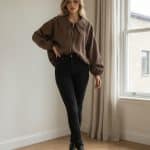 Mujer joven caminando en un interior luminoso junto a una ventana, vistiendo una chaqueta amplia marrón tipo bomber con cremallera mangas murciélago y zapatillas blancas.