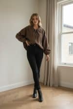 Mujer joven caminando en un interior luminoso junto a una ventana, vistiendo una chaqueta amplia marrón tipo bomber con cremallera mangas murciélago y zapatillas blancas.