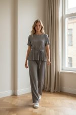 Conjunto femenino plisado gris compuesto por blusa de manga corta con cintura ajustable y pantalón palazzo, usado en interior junto a una ventana.