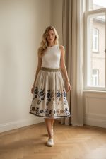 Falda midi color crema con bordado floral azul y dorado en el bajo, corte evasé y cintura elástica.