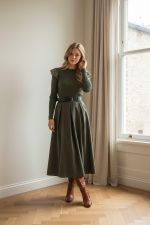 Falda midi verde de vuelo con cintura alta y cinturón negro, combinada con blusa beige y botines marrones.