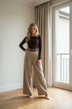 Mujer de pie junto a una ventana vistiendo pantalón palazzo beige de talle alto con cinturón, perneras amplias y largo completo, combinado con top marrón ajustado y zapatos de tacón claros.