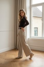 Mujer de pie junto a una ventana vistiendo pantalón palazzo beige de talle alto con cinturón, perneras amplias y largo completo, combinado con top marrón ajustado y zapatos de tacón claros.