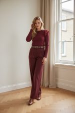 Mujer de pie junto a una ventana vistiendo pantalón palazzo burdeos de talle alto con cinturón, perneras amplias y largo completo, combinado con top marrón ajustado y zapatos de tacón claros.