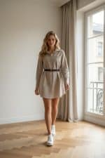 Mujer posando junto a una ventana con vestido camisero beige ceñido con cinturón negro y botines blancos.