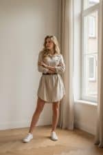 Mujer posando junto a una ventana con vestido camisero beige ceñido con cinturón negro y botines blancos.