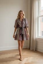 Mujer joven caminando en interior luminoso junto a una ventana, con vestido corto estampado de mangas abullonadas y cinturón negro.