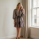 Mujer joven caminando en interior luminoso junto a una ventana, con vestido corto estampado de mangas abullonadas y cinturón negro.