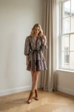 Mujer joven caminando en interior luminoso junto a una ventana, con vestido corto estampado de mangas abullonadas y cinturón negro.