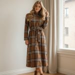 Vestido midi marrón de cuadros tipo tartán con cinturón del mismo tejido, manga larga y cuello redondo, combinado con botines marrones.